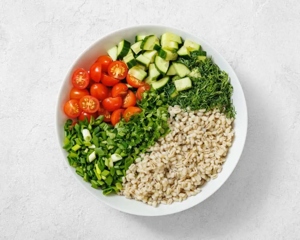A white bowl containing chopped cucumber, cherry tomatoes, fresh dill, green onions, and cooked barley arranged in separate sections.