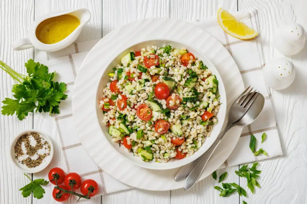 A bowl of barley salad with cherry tomatoes, cucumber, and parsley, surrounded by lemon wedge, dressing, herbs, salt, pepper, and cutlery on a white wooden table.