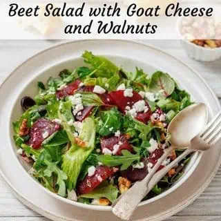 A salad with mixed greens, beets, feta, and walnuts on a plate. Beside it are a baguette, salt and pepper shakers, and a dish of jam.