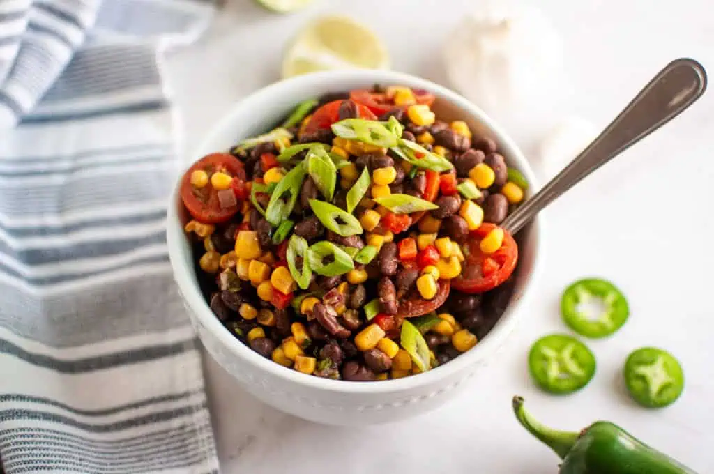 A white bowl filled with a salad of black beans, corn, cherry tomatoes, diced red pepper, and sliced green onions, with a spoon inside. Sliced jalapeños and a lime are nearby.
