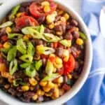 A bowl of black bean and corn salad topped with sliced cherry tomatoes and chopped green onions, placed on a white surface next to a blue napkin.
