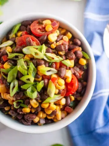 A bowl of black bean and corn salad topped with sliced cherry tomatoes and chopped green onions, placed on a white surface next to a blue napkin.