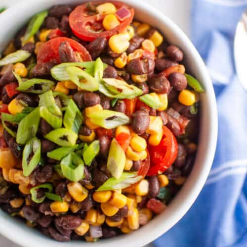 A bowl of black bean and corn salad topped with sliced cherry tomatoes and chopped green onions, placed on a white surface next to a blue napkin.