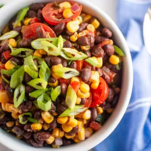 A bowl of black bean and corn salad topped with sliced cherry tomatoes and chopped green onions, placed on a white surface next to a blue napkin.