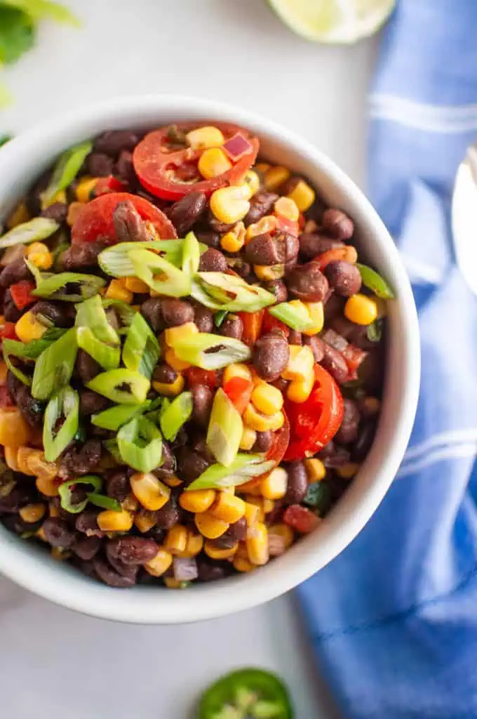 A bowl of black bean and corn salad topped with sliced cherry tomatoes and chopped green onions, placed on a white surface next to a blue napkin.
