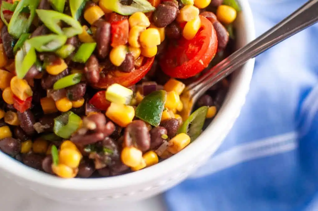 A close-up of a bowl filled with Black Bean & Corn Fiesta Salad, featuring black beans, corn, cherry tomatoes, and green onions, with a spoon inside.