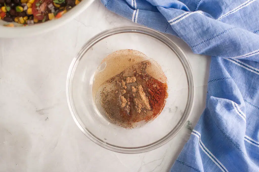 A glass bowl containing ground spices and liquid sits on a marble surface next to a blue kitchen towel. Part of another bowl with mixed vegetables is visible in the corner.