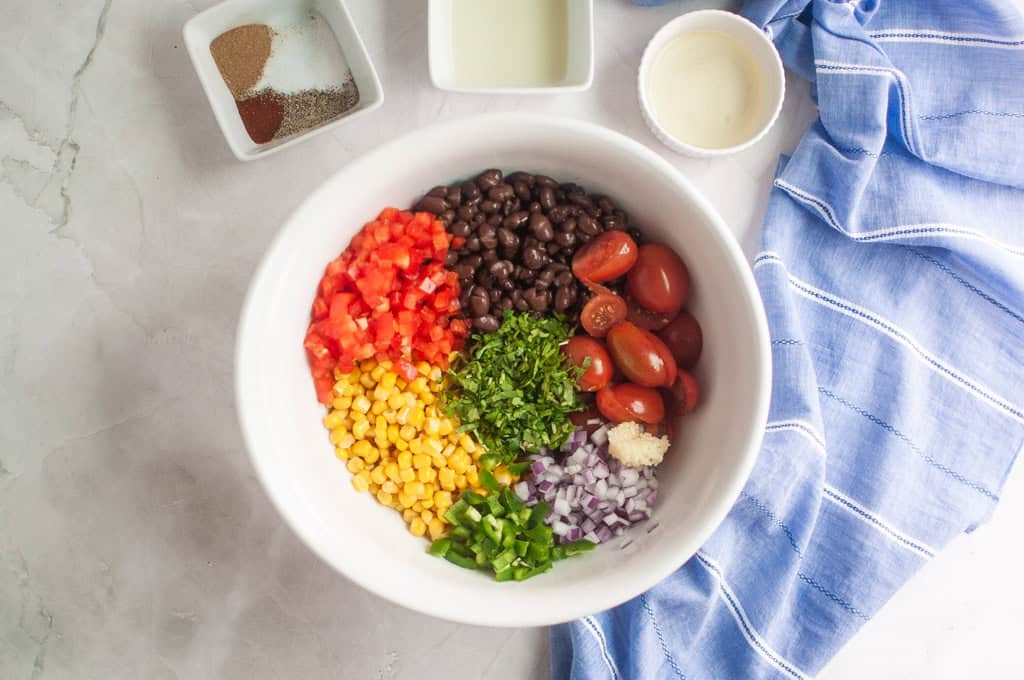 A white bowl contains black beans, diced tomatoes, corn, chopped cilantro, cherry tomatoes, chopped onion, jalape&ntilde;o, and minced garlic. Small bowls of spices and liquids are nearby.