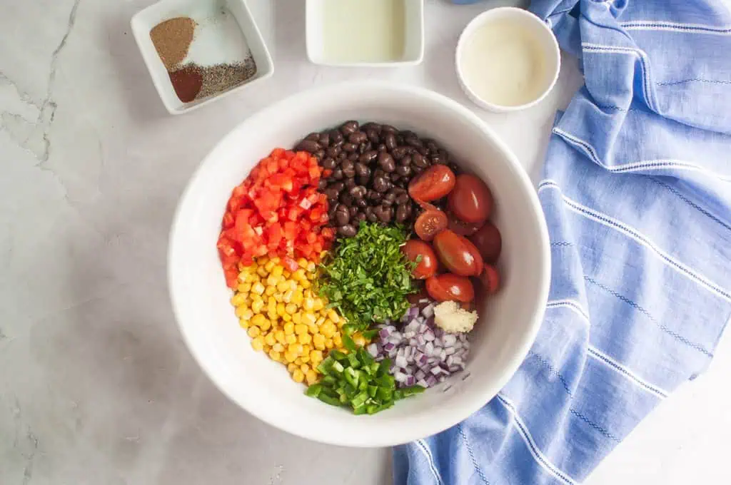 A white bowl contains black beans, diced tomatoes, corn, chopped cilantro, cherry tomatoes, chopped onion, jalapeño, and minced garlic. Small bowls of spices and liquids are nearby.