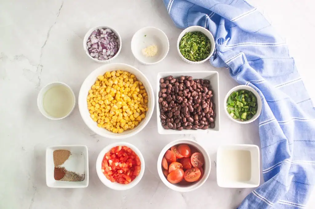 Ingredients for a salad or salsa arranged in bowls: corn, black beans, cherry tomatoes, red bell pepper, onion, jalapeño, cilantro, spices, lime juice, minced garlic, and a blue towel.