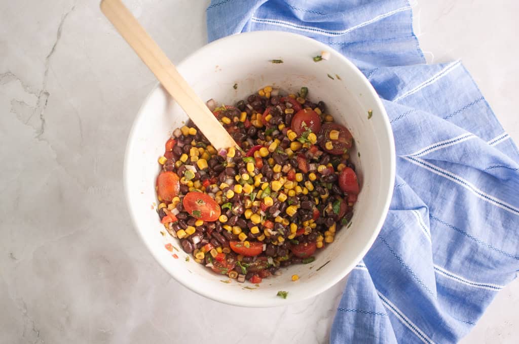 A white bowl with a mixture of black beans, corn, cherry tomatoes, and herbs, with a wooden spoon inside, next to a blue striped cloth on a light surface.