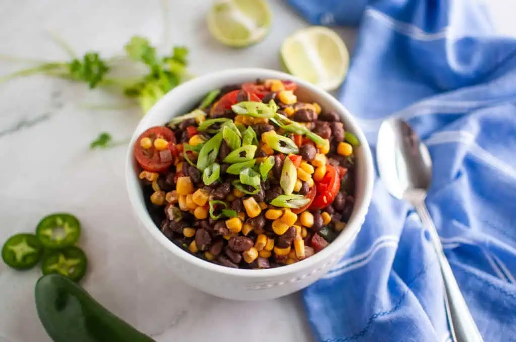 A bowl of black bean and corn salad topped with sliced green onions and cherry tomatoes, with lime halves, cilantro, jalapeño, and a blue cloth napkin nearby.