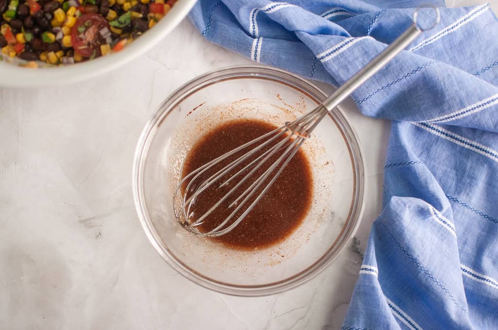 A glass bowl with a wire whisk mixing a brown liquid, next to a blue striped cloth and a bowl of colorful vegetables on a marble surface.