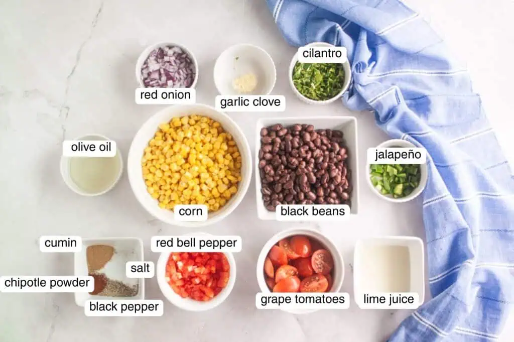 Ingredients for a black bean and corn salad arranged in bowls, including corn, black beans, tomatoes, red onion, red bell pepper, jalapeño, cilantro, garlic, spices, olive oil, and lime juice.