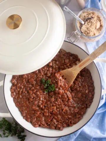 A white Dutch oven with baked beans topped with parsley sits on a marble surface next to a jar of brown sugar, a wooden spoon, and a blue cloth.