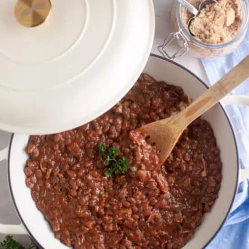 A white Dutch oven with baked beans topped with parsley sits on a marble surface next to a jar of brown sugar, a wooden spoon, and a blue cloth.