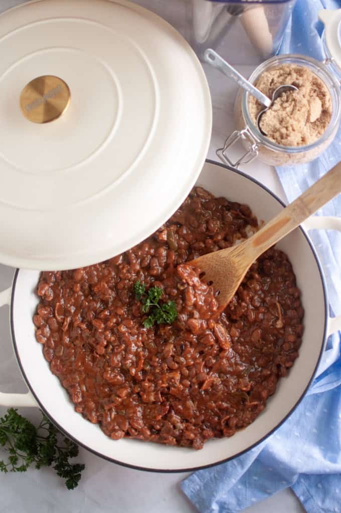 A white Dutch oven with baked beans topped with parsley sits on a marble surface next to a jar of brown sugar, a wooden spoon, and a blue cloth.