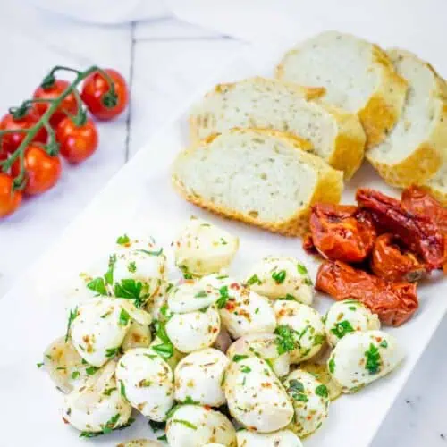 A rectangular white plate with sliced bread, marinated mozzarella balls with herbs, roasted tomatoes, and a vine of cherry tomatoes on the side.