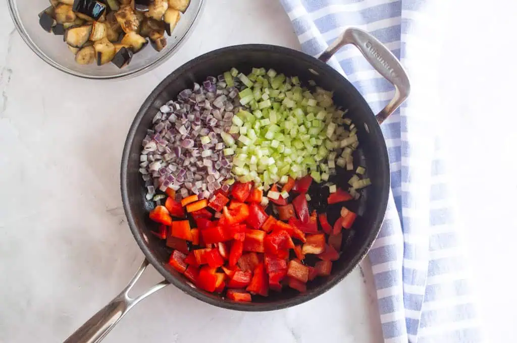 A skillet with diced red bell pepper, celery, and red onion, with a bowl of cooked eggplant and a striped towel beside it on a marble surface.