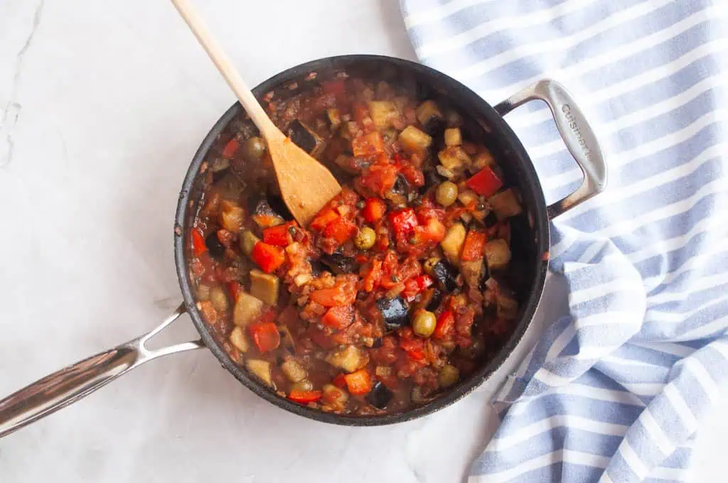A pan of vegetable stew with tomatoes, eggplant, and olives sits on a marble surface next to a blue-and-white striped towel; a wooden spoon rests in the pan.