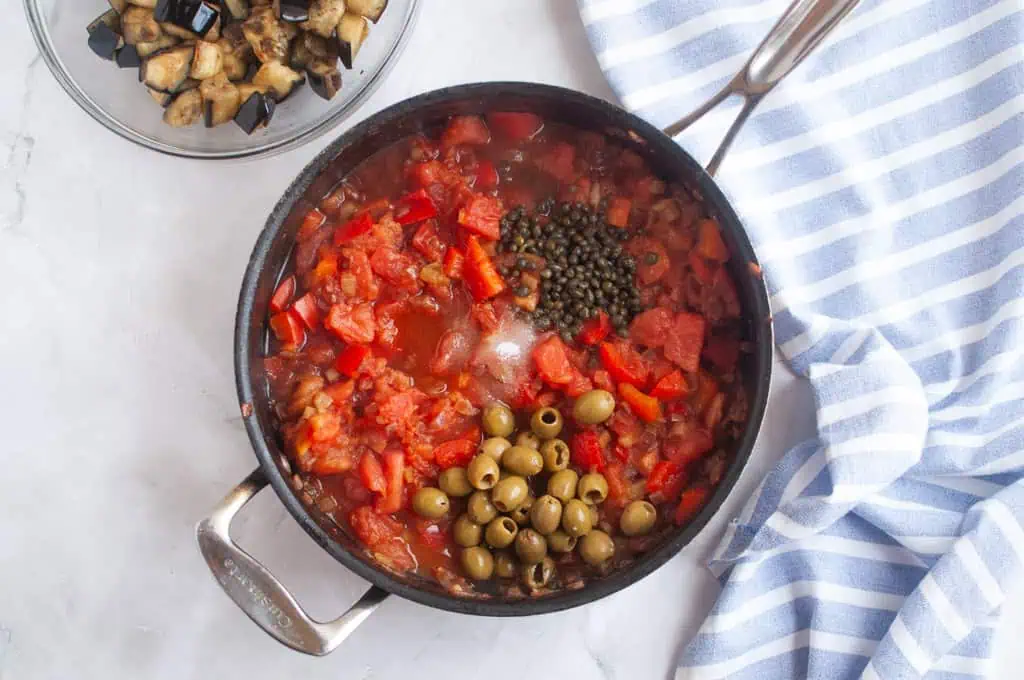 A pan filled with chopped tomatoes, green olives, capers, and vegetables sits on a counter next to a bowl of diced eggplant and a striped kitchen towel.