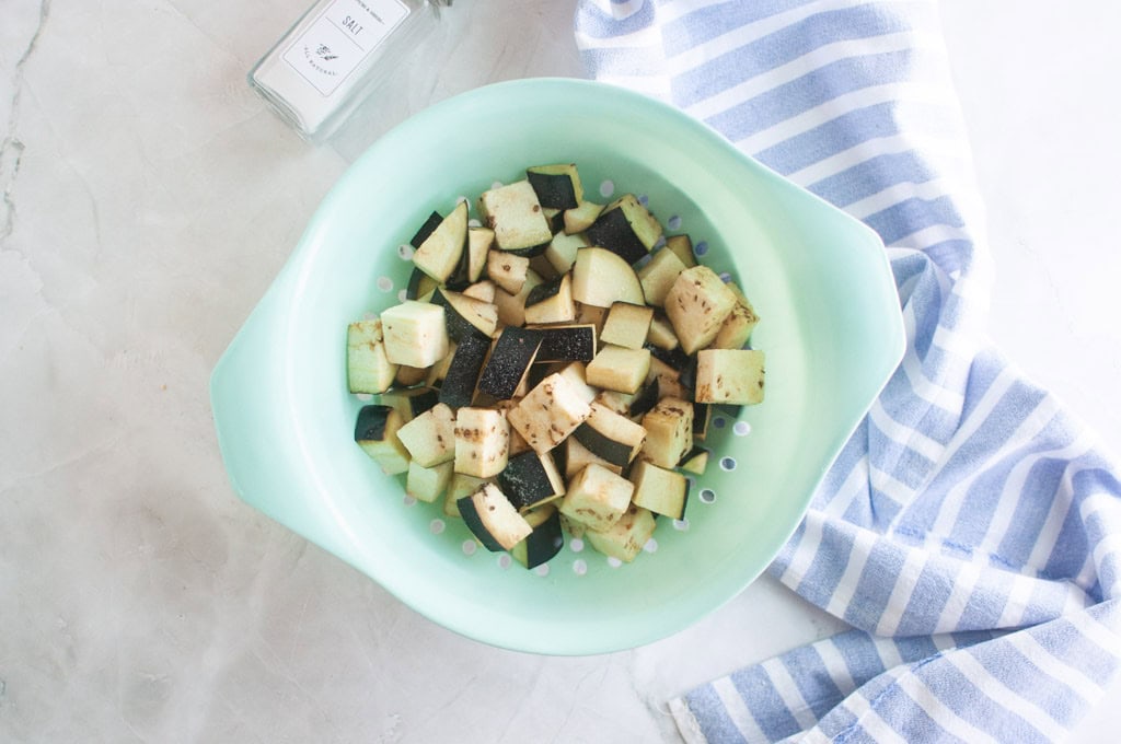 Cubed eggplant pieces in a light green colander on a marble surface, next to a blue and white striped cloth and a glass jar.