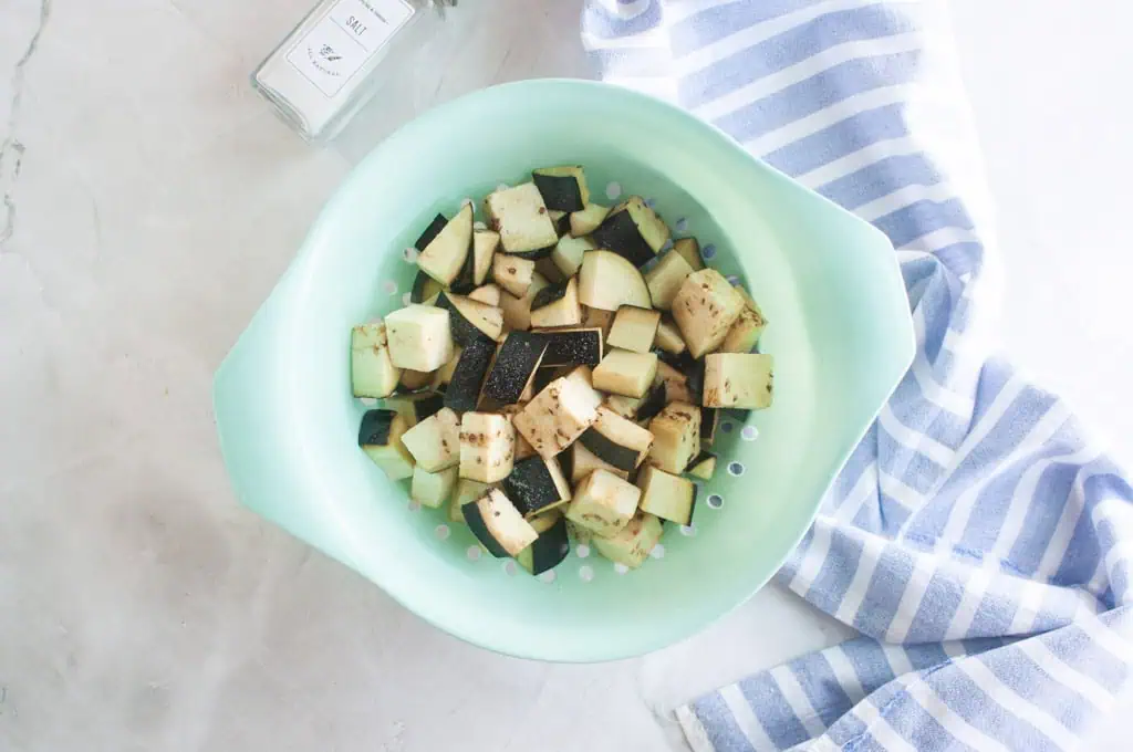 Cubed eggplant pieces in a light green colander, next to a salt container and a blue and white striped dish towel on a light surface.
