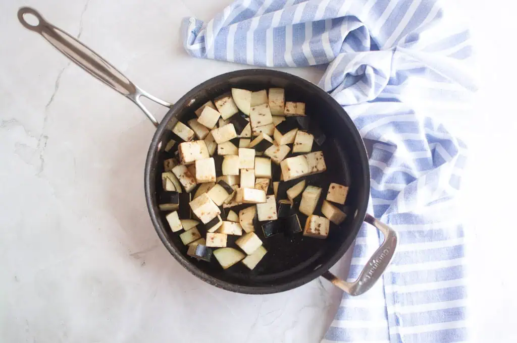 A black saucepan containing cubed eggplant sits on a light countertop next to a blue and white striped cloth.