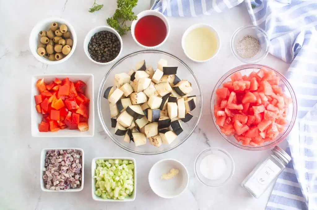 Chopped eggplant, tomatoes, red bell pepper, celery, shallots, garlic, olives, capers, and seasonings arranged in bowls on a white surface.