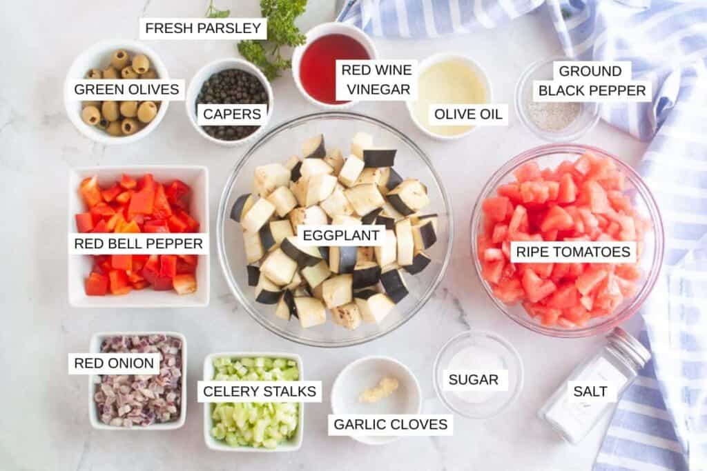 Ingredients for a recipe arranged in bowls on a white surface, including eggplant, tomatoes, peppers, onions, olives, capers, celery, garlic, parsley, and various seasonings.