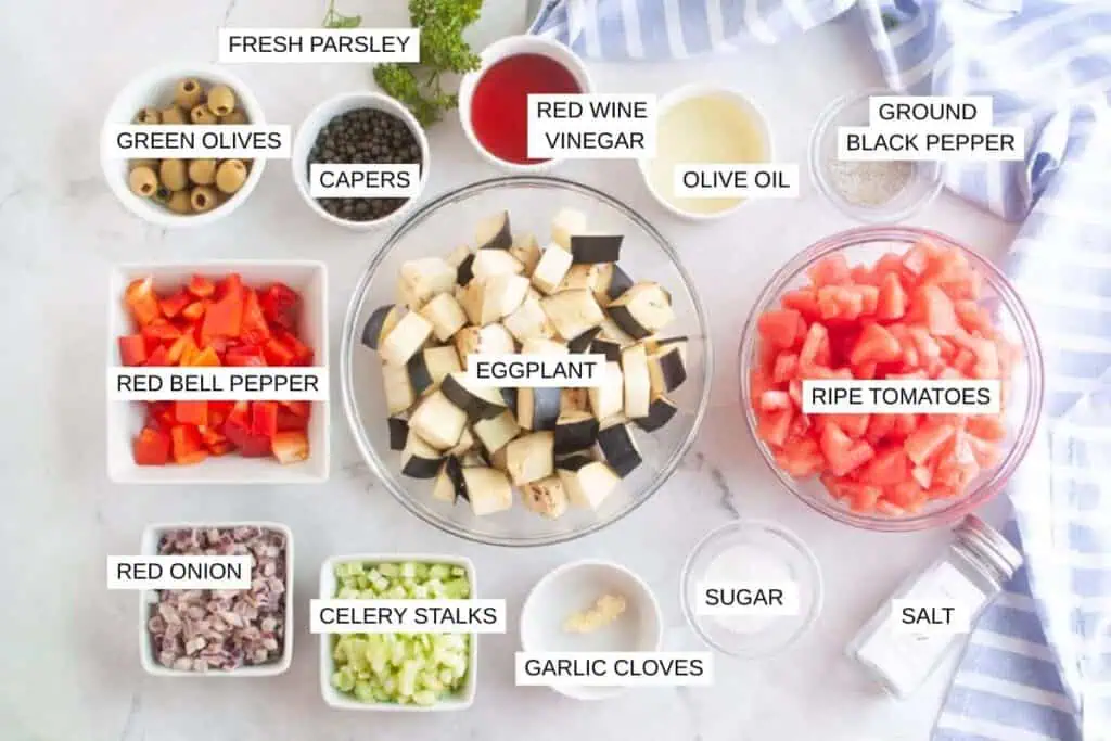 Ingredients for a recipe arranged in bowls on a white surface, including eggplant, tomatoes, peppers, onions, olives, capers, celery, garlic, parsley, and various seasonings.