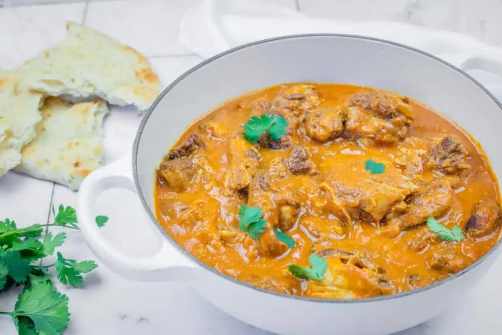 A white pot filled with chicken curry in orange sauce, garnished with cilantro, sits on a marble surface next to pieces of naan bread and fresh cilantro.