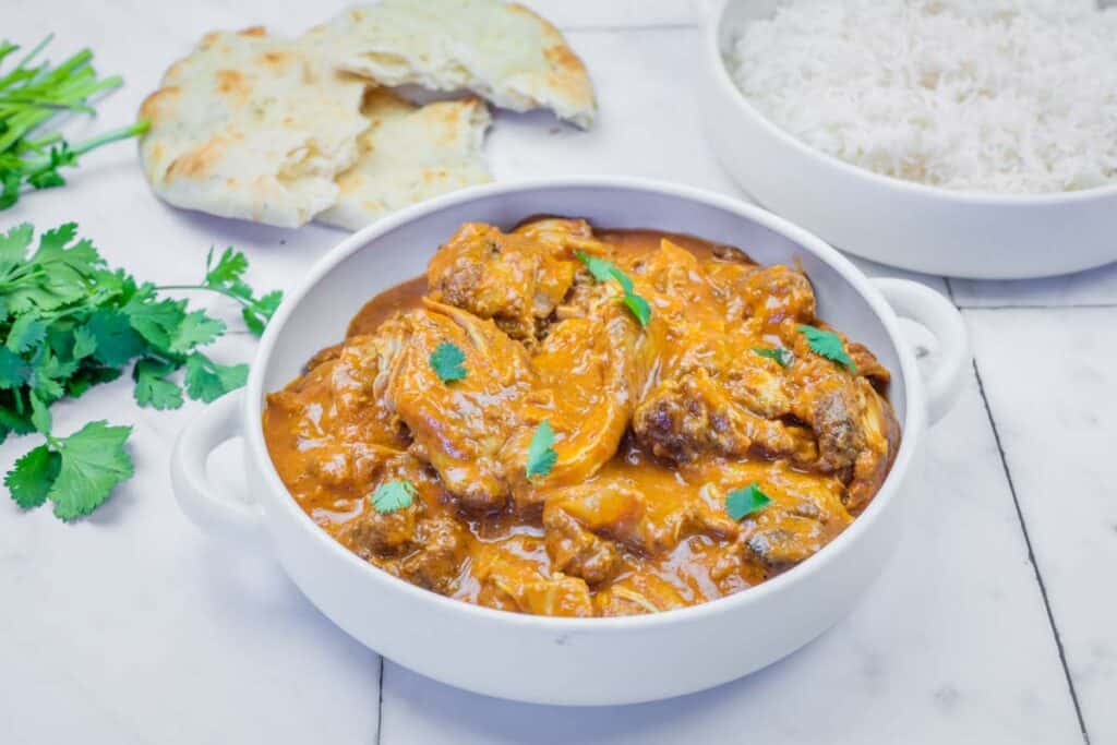 A white pot filled with chicken curry in orange sauce, garnished with cilantro, sits on a marble surface next to pieces of naan bread and fresh cilantro.