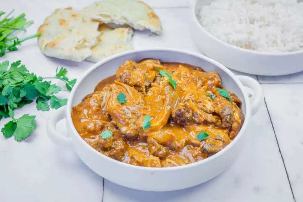A white pot filled with chicken curry in orange sauce, garnished with cilantro, sits on a marble surface next to pieces of naan bread and fresh cilantro.