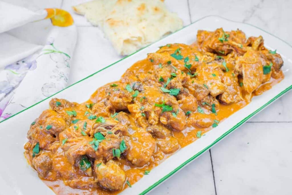 A rectangular white plate with chicken tikka masala garnished with chopped herbs, served on a marble surface with naan bread in the background.