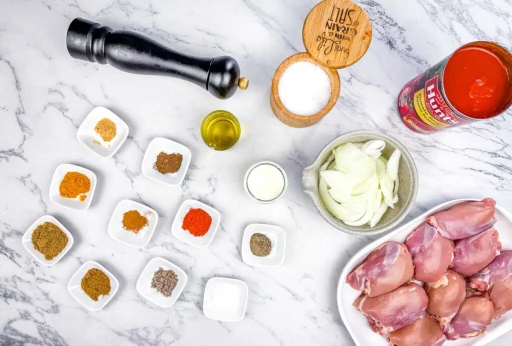 Ingredients for a chicken curry dish laid out on a marble surface, including raw chicken thighs, spices, sliced onions, olive oil, salt, pepper, cream, and canned tomatoes.