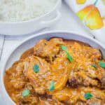 A bowl of chicken curry garnished with cilantro next to a plate of white rice, with a floral napkin in the background.
