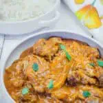 A bowl of chicken curry garnished with cilantro next to a plate of white rice, with a floral napkin in the background.