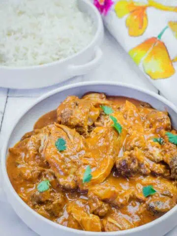 A bowl of chicken curry garnished with cilantro next to a plate of white rice, with a floral napkin in the background.