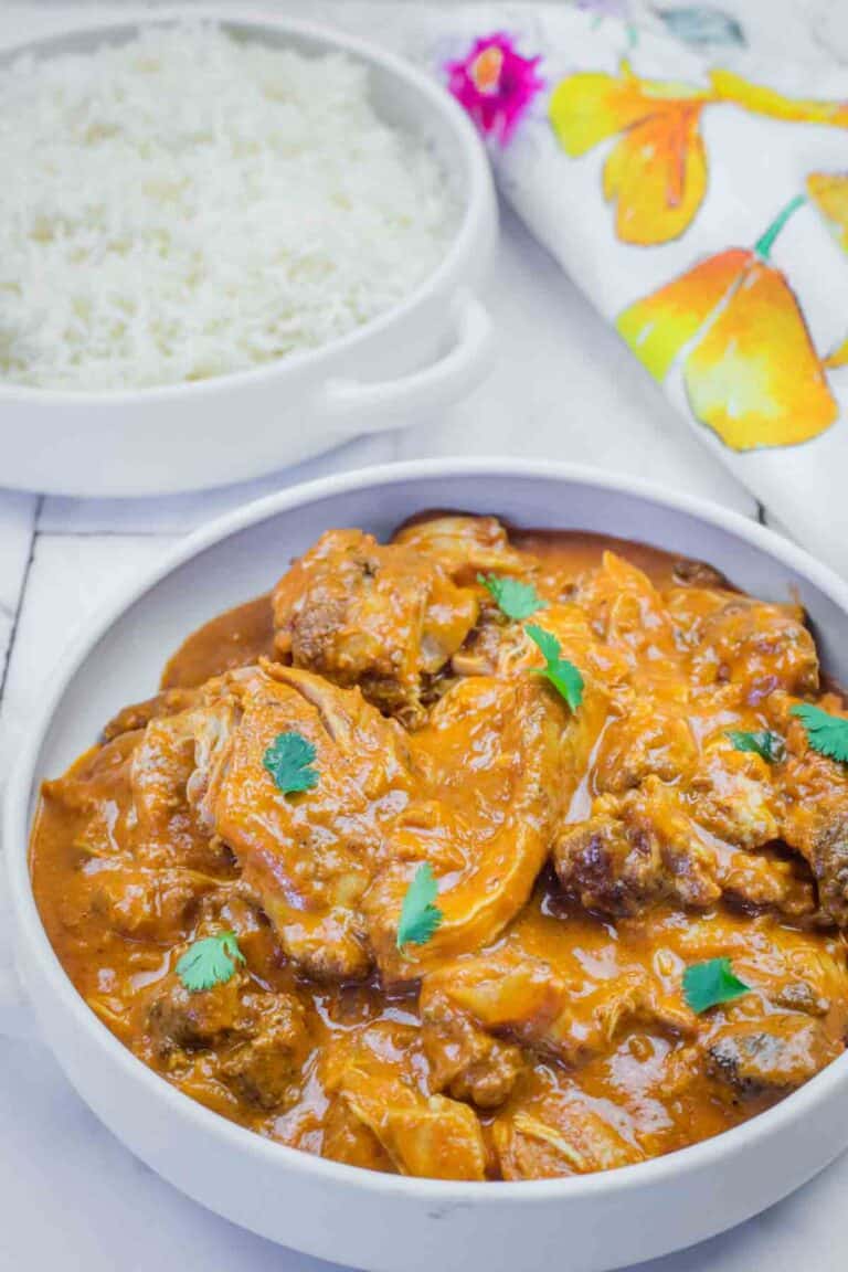 A bowl of chicken curry garnished with cilantro next to a plate of white rice, with a floral napkin in the background.