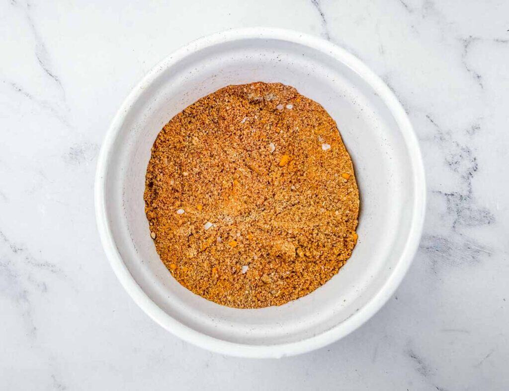 A white bowl filled with a mixed blend of orange and brown spices sits on a white marble surface.