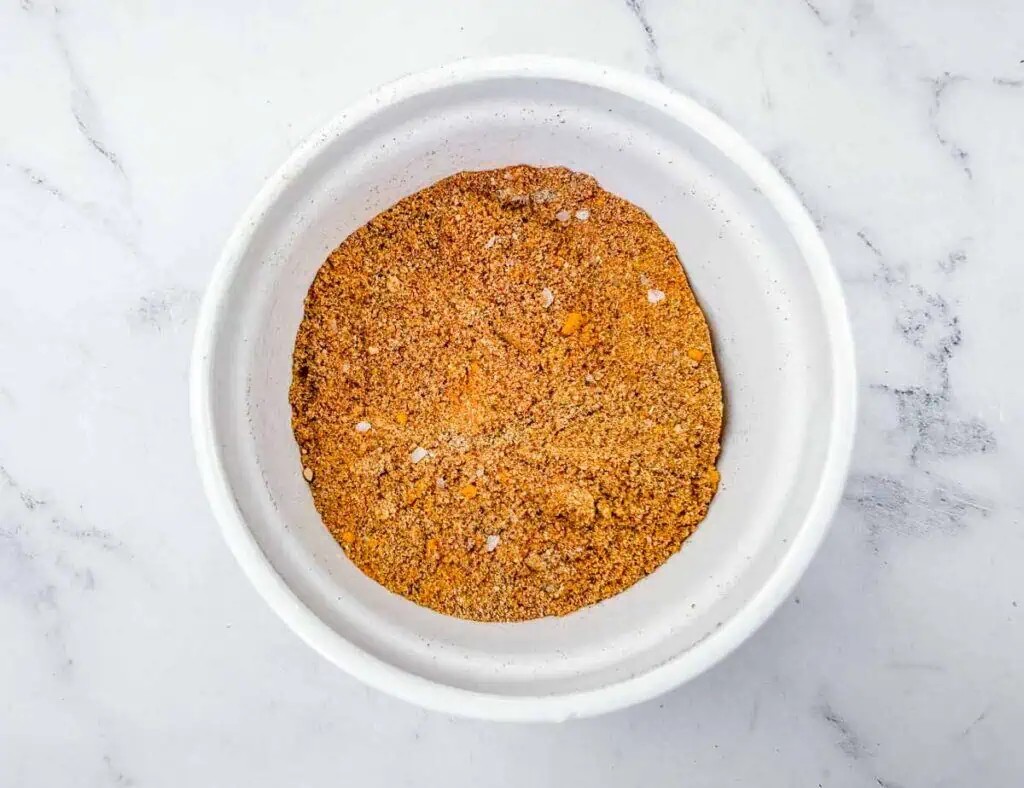 A white bowl filled with a mixed blend of orange and brown spices sits on a white marble surface.