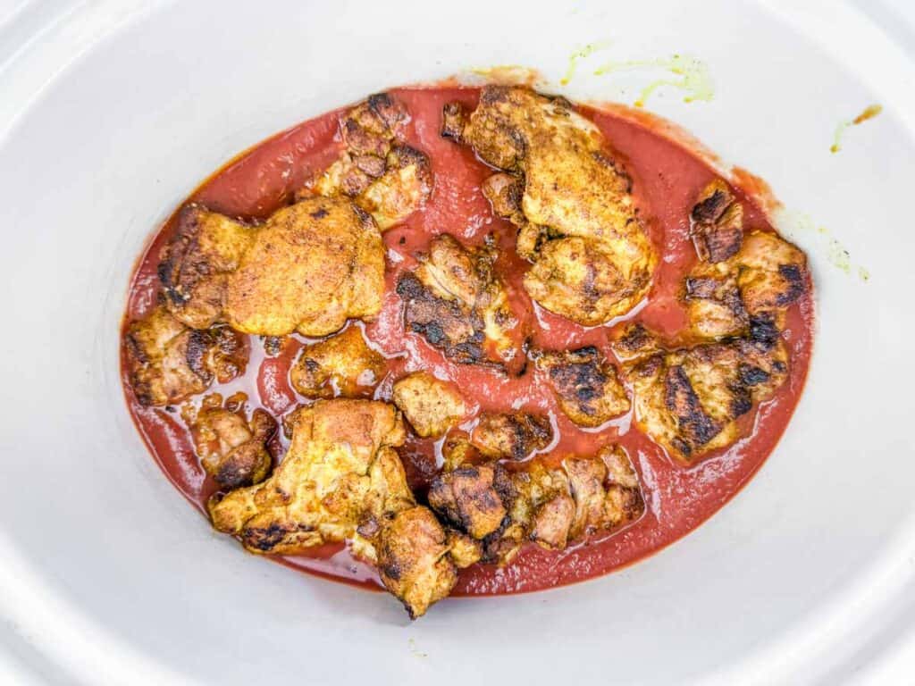 Seasoned, browned chicken pieces in tomato sauce inside a white slow cooker.