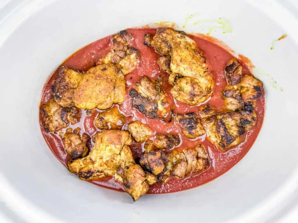 Seasoned, browned chicken pieces in tomato sauce inside a white slow cooker.