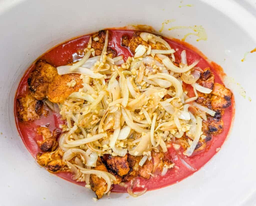 Cooked chunks of seasoned chicken, sliced onions, and garlic sit in tomato sauce inside a white slow cooker.