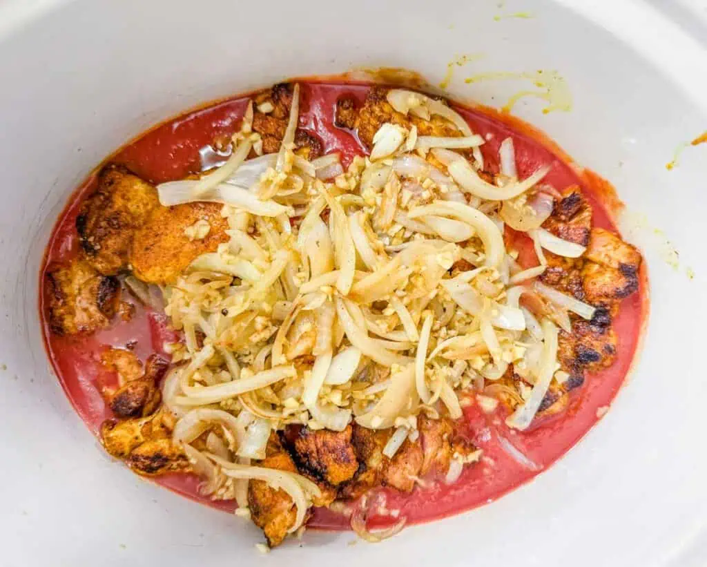 Cooked chunks of seasoned chicken, sliced onions, and garlic sit in tomato sauce inside a white slow cooker.