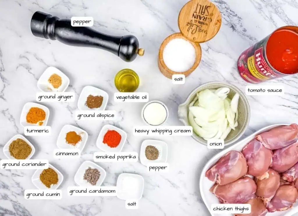 Labeled ingredients for a chicken recipe are arranged on a marble surface, including spices, chicken thighs, onion, tomato sauce, cream, oil, salt, and pepper.