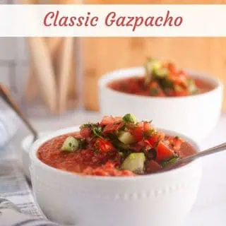 Two white bowls of classic gazpacho soup, garnished with chopped vegetables and herbs.