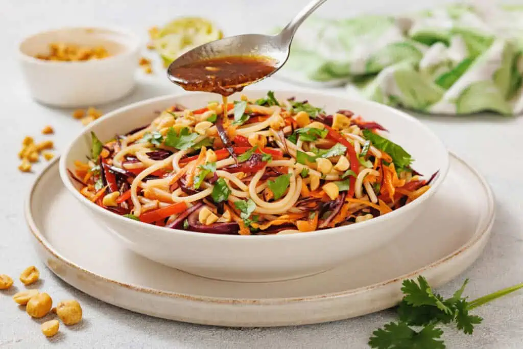 A bowl of noodle salad with shredded vegetables and peanuts is being topped with sauce from a spoon. A small dish of peanuts and a lime wedge are in the background.