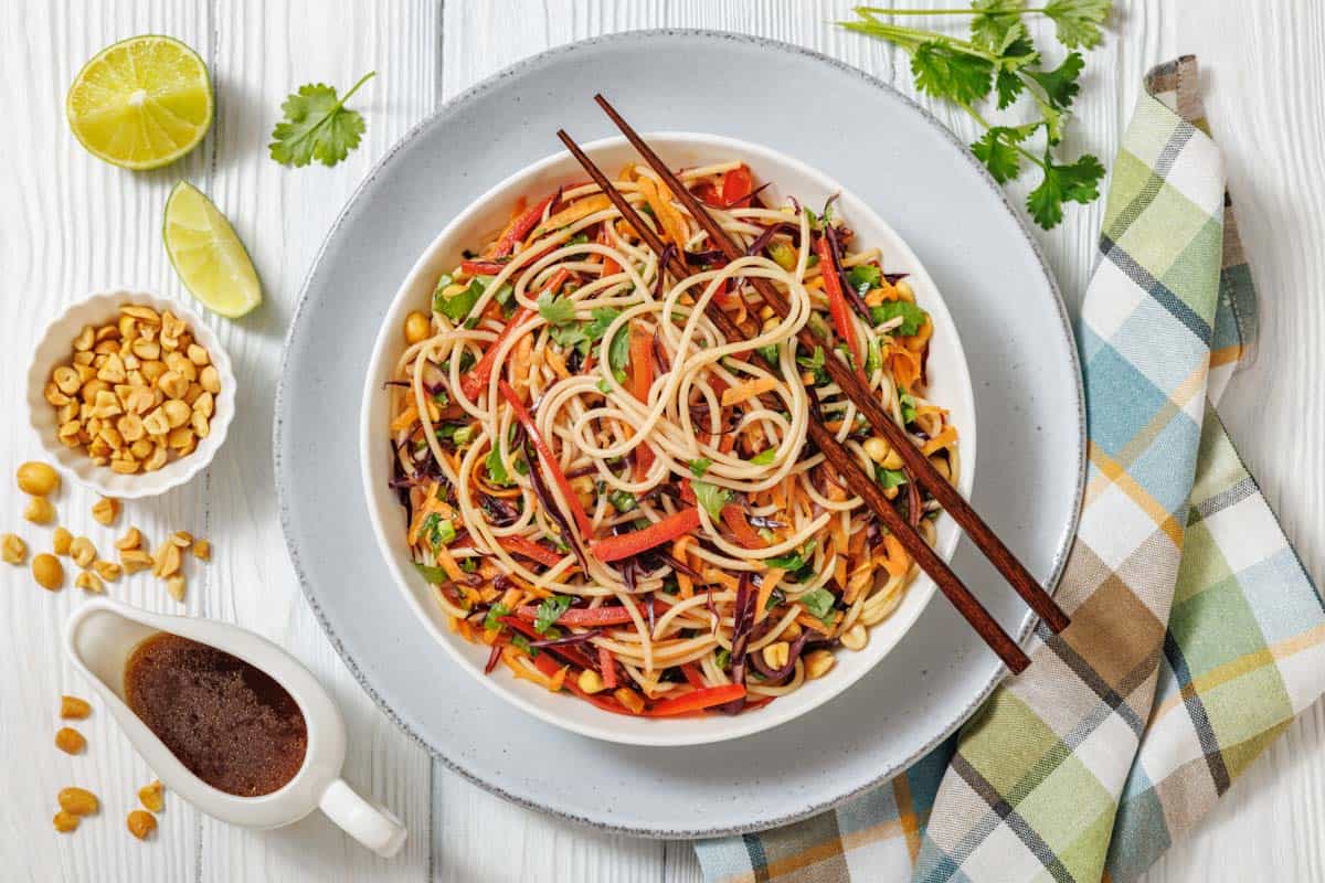 A bowl of Asian noodle salad with vegetables and chopsticks, surrounded by lime, peanuts, sauce, and fresh cilantro on a light wooden table with a plaid napkin.